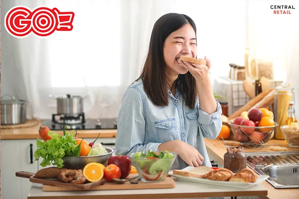 Một người phụ nữ trẻ đang mỉm cười và thưởng thức bánh mì gối trong không gian ấm cúng, thể hiện sự thoải mái