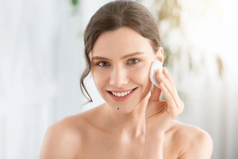 Một người đang dùng bông tẩy trang lau mặt, tránh vùng da vừa tẩy nốt ruồi
